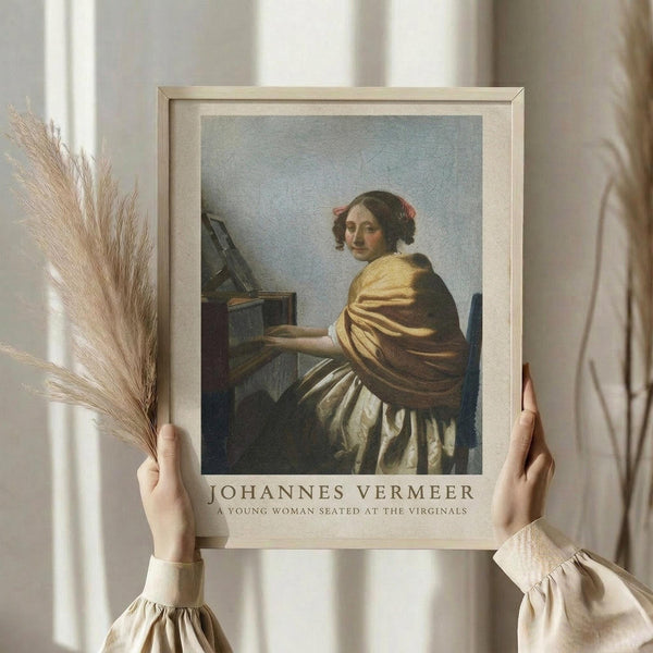 May include: Hands holding a framed art print of Vermeer's painting showing a young woman in golden yellow dress seated at virginals. Light wood frame with white matting, dried pampas grass stems visible on left side. Person wearing cream colored clothing with ruffled sleeves against soft neutral background with window light.
