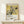 May include: Framed Japanese woodblock print poster featuring white hydrangea flowers with dark green leaves and a small black bird against a golden yellow background. The artwork displays vertical Japanese text and "KATSUSHIKA HOKUSAI" with dates 1822-1834 in the lower portion. Natural wood frame sits on light wooden furniture next to a small white wrapped gift with twine and evergreen sprig. Cream colored wall background creates minimalist styling.