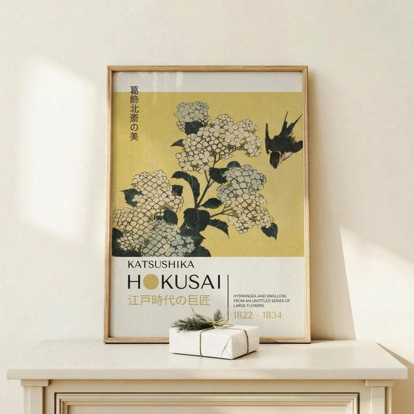 May include: Framed Japanese woodblock print poster featuring white hydrangea flowers with dark green leaves and a small black bird against a golden yellow background. The artwork displays vertical Japanese text and "KATSUSHIKA HOKUSAI" with dates 1822-1834 in the lower portion. Natural wood frame sits on light wooden furniture next to a small white wrapped gift with twine and evergreen sprig. Cream colored wall background creates minimalist styling.
