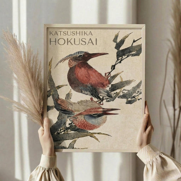 May include: Person in cream ruffled sleeves holding framed Japanese-style artwork featuring two colorful birds perched on green bamboo branches. The print shows reddish-brown and gray birds with long beaks in traditional woodblock print style against a cream background. Natural dried wheat stalks visible on left side, soft window lighting creates warm atmosphere.