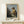 May include: Framed art print of a classical painting showing a young woman in golden yellow and white clothing seated at a musical instrument, displayed on a light wooden surface alongside a small white gift box with natural twine and greenery, against a cream colored wall in soft natural lighting.