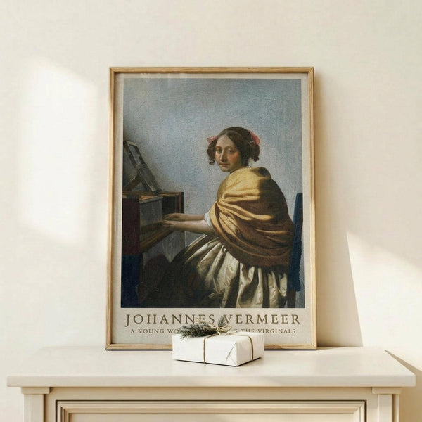 May include: Framed art print of a classical painting showing a young woman in golden yellow and white clothing seated at a musical instrument, displayed on a light wooden surface alongside a small white gift box with natural twine and greenery, against a cream colored wall in soft natural lighting.