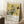 May include: Framed Japanese art print leaning against white wall beside wooden side table with coffee cup. Print features white and gray hydrangea flowers with dark leaves and flying bird on golden yellow background. Japanese text at top, "KATSUSHIKA HOKUSAI" and dates "1822-1834" at bottom. Thin natural wood frame, positioned on white textured rug next to cream blanket.