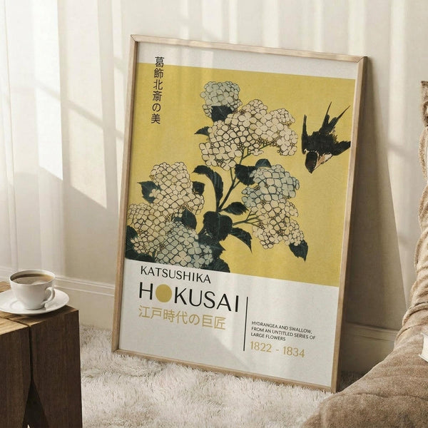 May include: Framed Japanese art print leaning against white wall beside wooden side table with coffee cup. Print features white and gray hydrangea flowers with dark leaves and flying bird on golden yellow background. Japanese text at top, "KATSUSHIKA HOKUSAI" and dates "1822-1834" at bottom. Thin natural wood frame, positioned on white textured rug next to cream blanket.