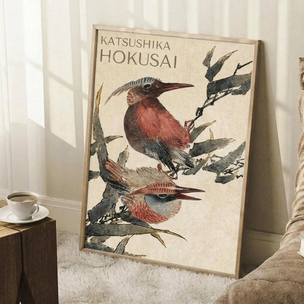 May include: Framed vintage-style Japanese woodblock print featuring two colorful birds perched on bamboo branches, displayed in a cozy room setting with white walls, wooden side table with coffee cup, and beige textiles. The artwork shows detailed illustrations in muted earth tones with the artist's name at the top.