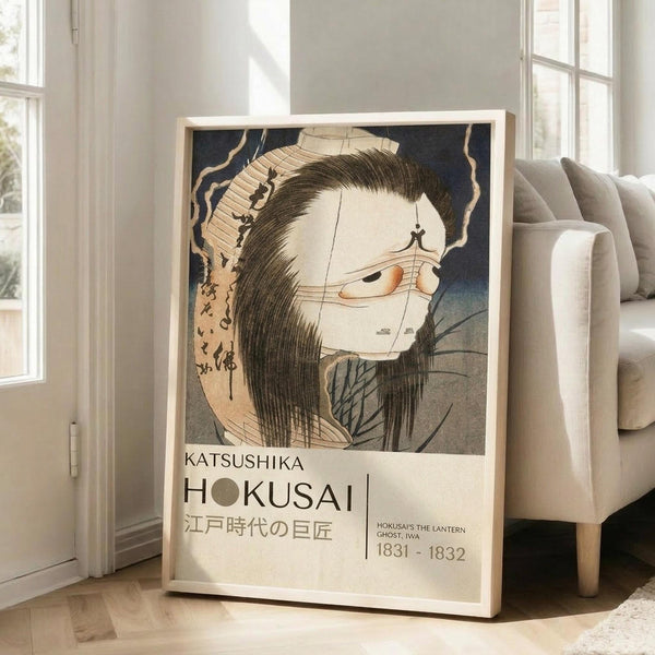 May include: A framed Japanese woodblock print featuring a ghostly figure with long dark hair, pale skin, and an orange eye, displayed on wooden flooring next to a beige upholstered chair. The artwork shows traditional ukiyo-e style with Japanese text and includes exhibition-style labeling with "HOKUSAI" and dates "1831-1832". Natural lighting streams through white-framed windows in the bright, minimalist room setting.