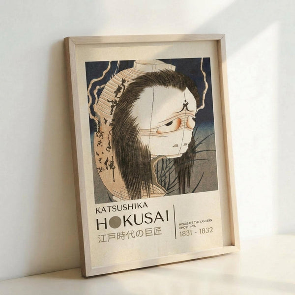 May include: Framed poster print featuring a Japanese woodblock artwork of a ghostly figure with long black hair, pale white face, and traditional clothing against a dark blue background. The print shows museum-style text below reading "KATSUSHIKA HOKUSAI" with Japanese characters and dates "1831-1832." The artwork is displayed in a light wooden frame, leaning against a white wall on what appears to be a light surface.