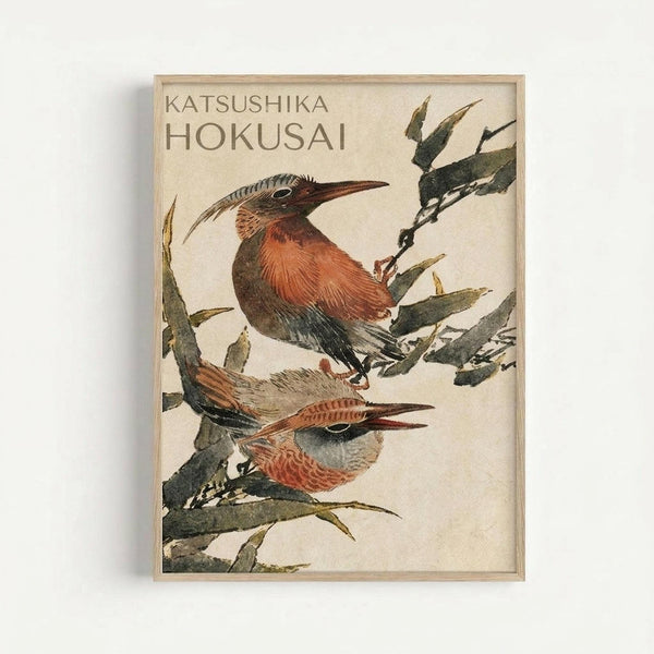 A framed Japanese woodblock print featuring two birds with reddish-brown plumage and long beaks perched among green bamboo or reed plants. The artwork displays traditional ukiyo-e style with muted earth tones against a cream background. Text reading "KATSUSHIKA HOKUSAI" appears in the upper left corner. The print is displayed in a light wooden frame against a white wall.