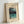 May include: Framed vintage Japanese woodblock print poster showing a serene lake scene with snow-capped mountains, tall trees with drooping branches, and turquoise water reflecting the landscape. The artwork features soft blues, greens, and coral tones in traditional ukiyo-e style. Light wood frame sits against white wall with natural lighting creating subtle shadows.