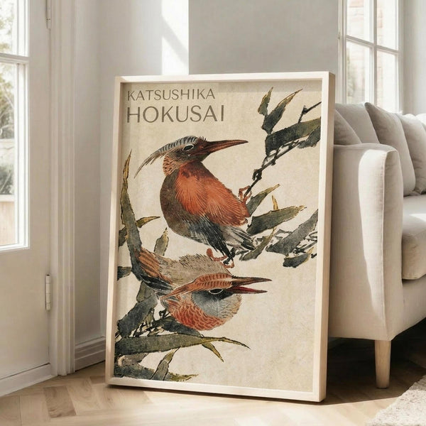 May include: A framed Japanese woodblock print featuring two russet-colored birds with long curved beaks perched on green bamboo branches against a cream background. The artwork displays traditional ukiyo-e style with visible text at the top. The light wood frame leans against a white wall next to a beige upholstered chair in a bright, naturally lit room with white window frames.
