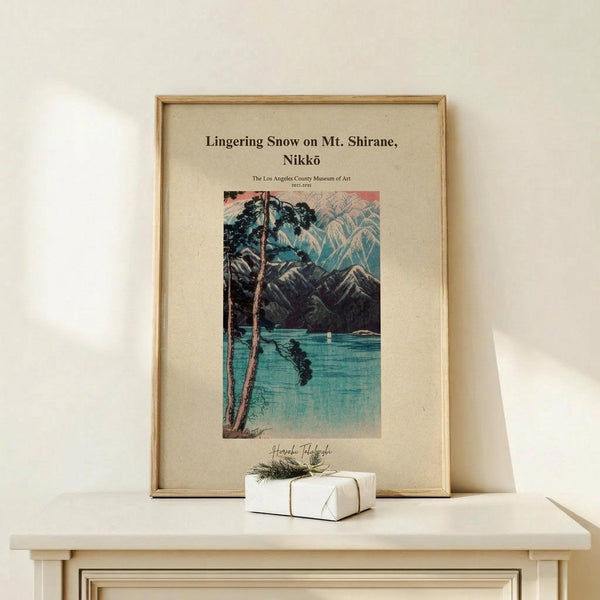 May include: A framed vintage Japanese woodblock print showing a serene lake scene with snow-capped mountains, pine trees, and turquoise water reflections. The artwork features muted teal, pink, and gray tones in a beige mat with thin wooden frame. Displayed on a light wood surface beside a small white wrapped gift with twine and greenery sprig against a cream wall.