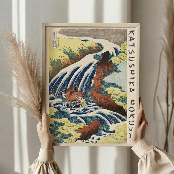 May include: Hands holding a framed Japanese woodblock print featuring a dramatic waterfall cascading down red-orange rocks with small figures below, displayed against white curtains with dried pampas grass visible to the left. The artwork shows traditional ukiyo-e style with blue water, green foliage, and vertical text along the right border.
