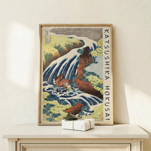 May include: A framed Japanese woodblock print featuring a dramatic waterfall cascading over rocky cliffs with small figures below, displayed on a light wood console table. The artwork shows traditional ukiyo-e style with blue water, orange-brown rocks, and green foliage against a gray sky. A small white wrapped gift sits in front of the natural wood frame on the minimal, cream-colored surface.