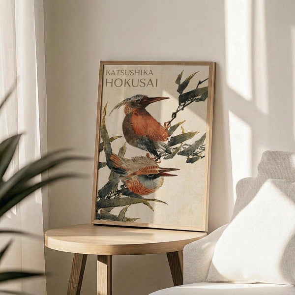 May include: Framed vintage Japanese bird artwork displayed on round wooden side table next to beige upholstered chair. Print features two colorful birds with orange-red breasts and long curved beaks perched among green foliage against cream background. Natural lighting creates soft shadows across neutral-toned room with plant visible in corner.