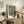 May include: Framed poster featuring a black cat with white eyes sitting beneath a decorative lantern and Japanese character on brown textured background, displayed on rustic wooden sideboard alongside fern plant, books, and ceramic vase in neutral-toned living room with beige furniture and walls