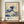 May include: A framed reproduction of Hokusai's famous Great Wave artwork displayed on a wooden sideboard against cream walls with white trim. The print shows the iconic blue and white wave with Mount Fuji in the background, featuring exhibition-style text below reading "Katsushika Hokusai" and "1830-1833." A small white ceramic vase and stack of books sit beside the brown wooden frame.