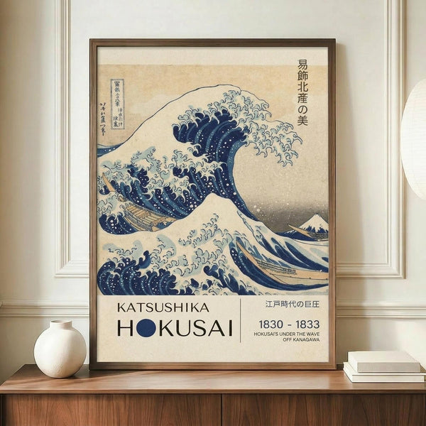 May include: A framed reproduction of Hokusai's famous Great Wave artwork displayed on a wooden sideboard against cream walls with white trim. The print shows the iconic blue and white wave with Mount Fuji in the background, featuring exhibition-style text below reading "Katsushika Hokusai" and "1830-1833." A small white ceramic vase and stack of books sit beside the brown wooden frame.