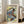 May include: Framed Japanese woodblock print artwork leaning against white wall next to beige sofa. The poster features a dramatic waterfall scene with cascading white water, orange-brown rocky cliffs, and small figures below. Traditional ukiyo-e style with blue, white, yellow and earth tones. Vertical text on cream background reads "KATSUSHIKA HOKUSAI" along the right edge. Natural lighting from nearby windows creates soft shadows on hardwood floor.
