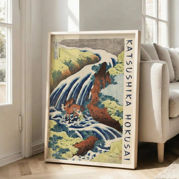 May include: Framed Japanese woodblock print artwork leaning against white wall next to beige sofa. The poster features a dramatic waterfall scene with cascading white water, orange-brown rocky cliffs, and small figures below. Traditional ukiyo-e style with blue, white, yellow and earth tones. Vertical text on cream background reads "KATSUSHIKA HOKUSAI" along the right edge. Natural lighting from nearby windows creates soft shadows on hardwood floor.