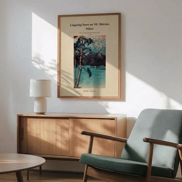 May include: Japanese woodblock print poster in wooden frame showing snow-covered mountain landscape with lake and pine trees in blues and greens. Displayed above mid-century modern wooden credenza with white ceramic lamp. Mint green upholstered armchair with wooden frame sits nearby. Warm sunlight creates shadows across white wall in minimalist living room setting.