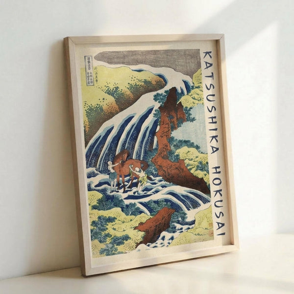 May include: Framed Japanese woodblock print reproduction showing a dramatic waterfall cascading down reddish-brown rocky cliffs with small figures at the base. The artwork features traditional ukiyo-e style with blue and white water, yellow-green foliage, and gray sky. Text reading "KATSUSHIKA HOKUSAI" runs vertically along the right edge. The print is displayed in a light wood frame leaning against a white wall.