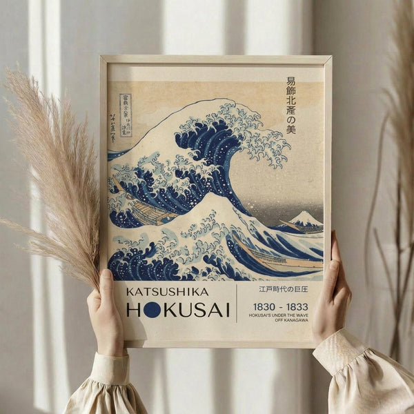May include: Hands holding a framed reproduction of Hokusai's famous Great Wave woodblock print in a light wooden frame. The artwork shows a large blue and white wave with Mount Fuji in the background, with exhibition-style text below reading "Katsushika Hokusai 1830-1833." Dried pampas grass sits beside the frame against a neutral beige backdrop with soft natural lighting.