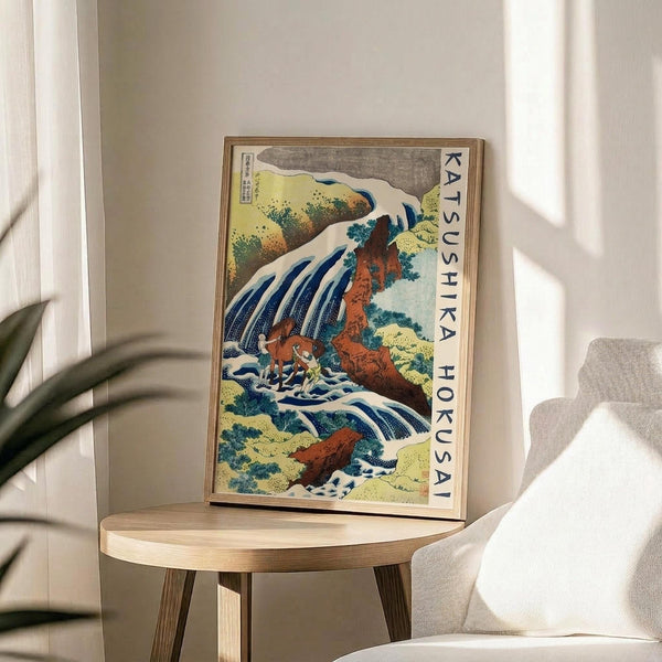 May include: Japanese woodblock print artwork in wooden frame showing a dramatic waterfall cascading over orange-red cliffs with blue and white water, surrounded by green foliage. The framed print sits on a light wood round side table next to a beige upholstered chair, with a potted plant visible in soft natural lighting from nearby windows.