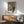 May include: Framed poster featuring a black cat character with wide eyes sitting beneath a white paper lantern with Japanese text, displayed in a mid-century modern living room with sage green upholstered chair, wood sideboard, white ceramic lamp, and natural sunlight casting shadows on white walls.
