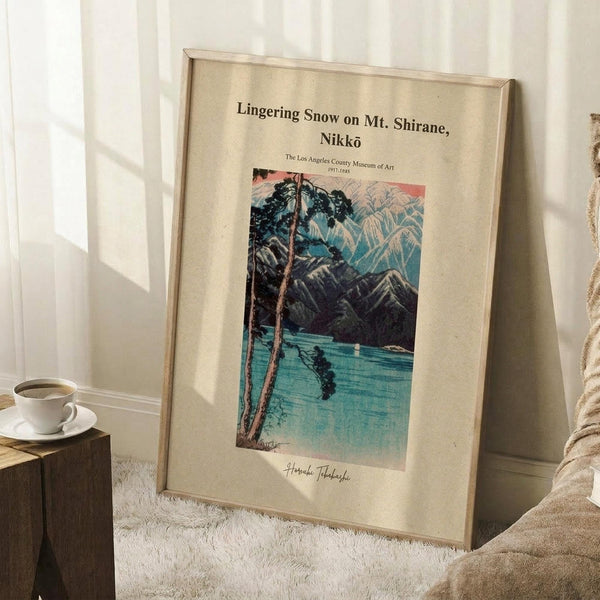 May include: Framed vintage Japanese woodblock print poster leaning against white wall corner on white textured rug. Artwork shows snowy mountain landscape with lake reflection in blue and pink tones, bare tree branches in foreground. Light wood frame with cream matting. Small wooden block with white coffee cup nearby. Soft natural lighting creates shadows on wall.