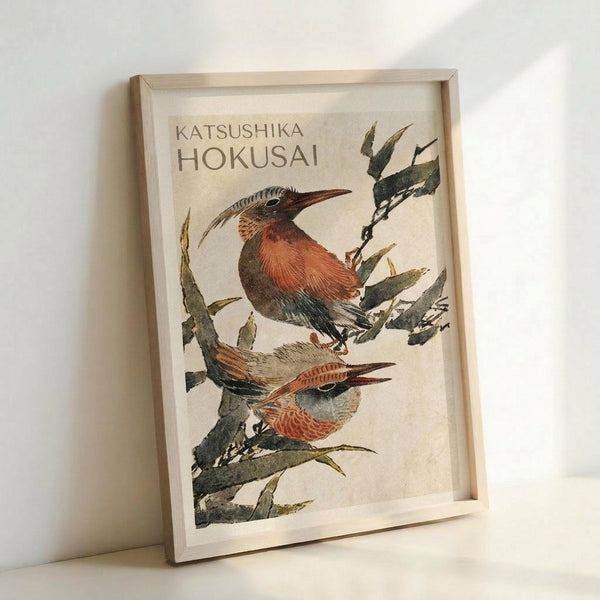 May include: Framed Japanese woodblock print artwork showing two colorful birds perched on green bamboo branches against a cream background. The birds have reddish-orange breasts, long beaks, and detailed feather patterns. Light wooden frame displayed leaning against white wall with soft natural lighting creating subtle shadows.