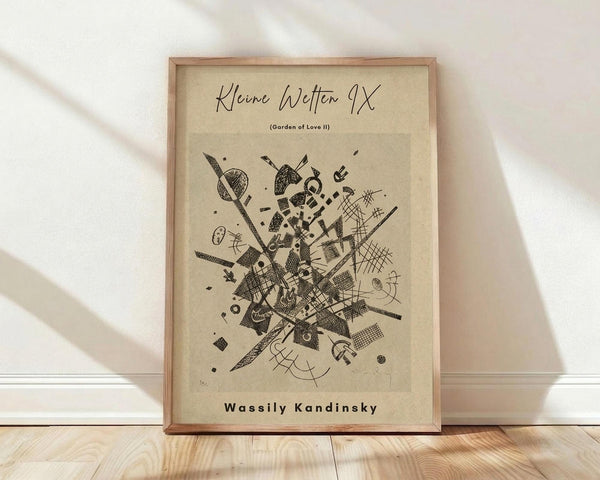 May include: Framed abstract art print leaning against white wall on light wood floor. Beige paper background with black geometric shapes, lines, circles, and crosshatched patterns in explosive radiating composition. Artist name "Wassily Kandinsky" at bottom. Natural wood frame with visible grain. Soft window light creates diagonal shadows across white textured wall.
