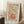 May include: Framed poster with retro-style illustration of an orange beer mug with colorful flowers and foliage sprouting from it, featuring text "Less Attitude More Gratitude" in black letters. The artwork uses warm autumn colors - oranges, browns, greens, and purples in a vintage folk art style. The poster leans against a white wall corner next to a wooden side table with coffee cup and saucer, positioned on a white textured rug.