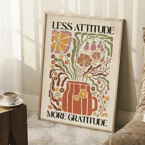 May include: Framed poster with retro-style illustration of an orange beer mug with colorful flowers and foliage sprouting from it, featuring text "Less Attitude More Gratitude" in black letters. The artwork uses warm autumn colors - oranges, browns, greens, and purples in a vintage folk art style. The poster leans against a white wall corner next to a wooden side table with coffee cup and saucer, positioned on a white textured rug.