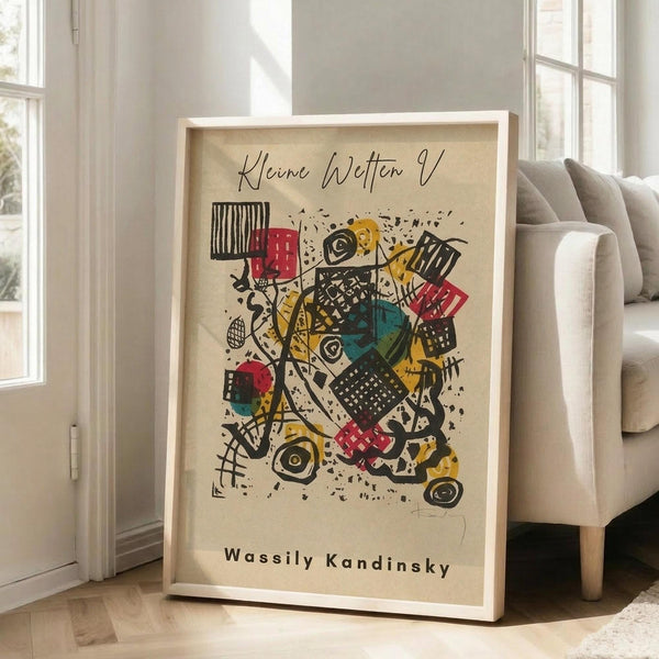 May include: Abstract art poster with colorful geometric shapes, lines, and patterns in yellow, red, teal, and black on cream background, displayed in light wood frame leaning against white wall next to beige couch in bright room with large windows and natural lighting on hardwood floors.