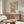 May include: Vintage-style poster with large orange ceramic vase and colorful floral arrangement, displayed above wooden mid-century credenza in modern living room. White sculptural chair and glass coffee table visible. Poster features retro botanical illustration in warm autumn tones of orange, brown, and green against cream background in thin wooden frame.