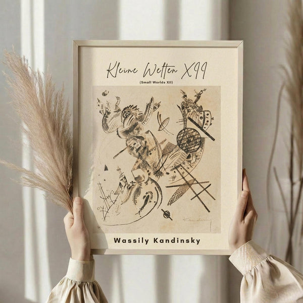 May include: Person in cream ruffle-sleeved top holding framed abstract art print in light wooden frame. The artwork features black line drawings with geometric shapes, organic forms, and cross-hatched patterns on aged beige paper. Dried pampas grass stems visible on left side. Soft natural lighting with white curtains in background creating neutral, minimalist home decor scene.