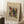 May include: Framed abstract art print leaning against white wall corner on textured rug. Artwork features colorful geometric shapes, patterns and forms in yellow, red, green, and black with "Wassily Kandinsky" text below. Natural wood frame with cream matting. Coffee cup on wooden block nearby, cozy room setting with soft natural lighting.
