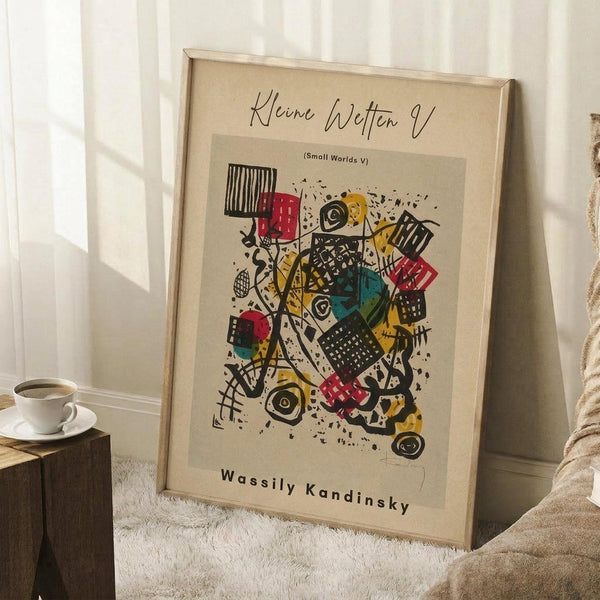 May include: Framed abstract art print leaning against white wall corner on textured rug. Artwork features colorful geometric shapes, patterns and forms in yellow, red, green, and black with "Wassily Kandinsky" text below. Natural wood frame with cream matting. Coffee cup on wooden block nearby, cozy room setting with soft natural lighting.