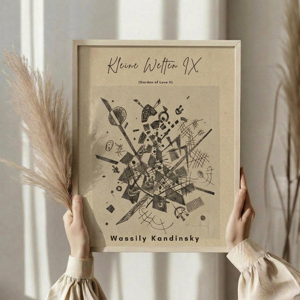 May include: Hands holding a framed abstract art print featuring geometric black line drawings and shapes on beige paper. The artwork shows intersecting lines, dots, crosshatching patterns, and angular forms in a dynamic composition. Person wears cream-colored ruffled sleeves and holds dried wheat stalks, photographed against soft white curtains in natural lighting.