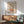 May include: Framed vintage-style poster with earth-toned botanical illustration featuring a wooden mug with steam swirls, butterfly, flowers, and text reading "Less Attitude More Gratitude." Displayed above a mid-century modern wood credenza with ceramic lamp, next to a sage green upholstered armchair with wooden frame. Natural sunlight creates geometric shadows on white wall, creating warm, cozy living room atmosphere.