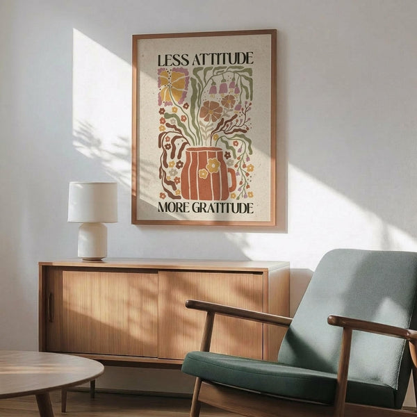 May include: Framed vintage-style poster with earth-toned botanical illustration featuring a wooden mug with steam swirls, butterfly, flowers, and text reading "Less Attitude More Gratitude." Displayed above a mid-century modern wood credenza with ceramic lamp, next to a sage green upholstered armchair with wooden frame. Natural sunlight creates geometric shadows on white wall, creating warm, cozy living room atmosphere.
