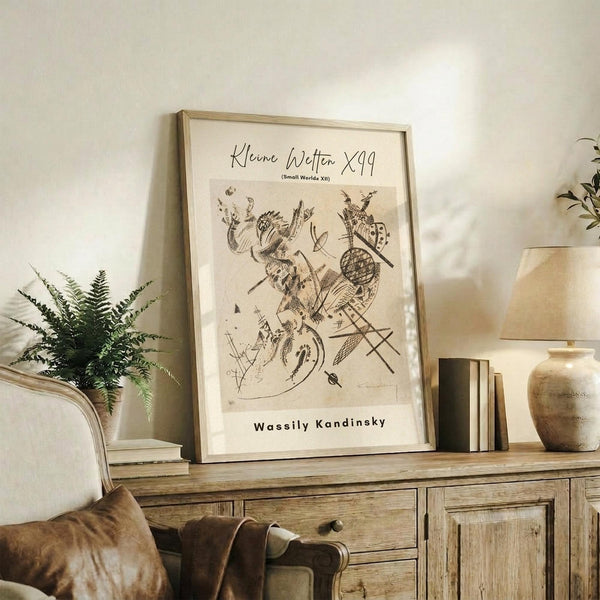 May include: Abstract art poster with intricate black line drawings featuring organic and geometric shapes, displayed in light wood frame on rustic wooden sideboard. Room scene shows beige upholstered chair, fern plant, ceramic vase, stacked books, and warm neutral lighting against cream walls.