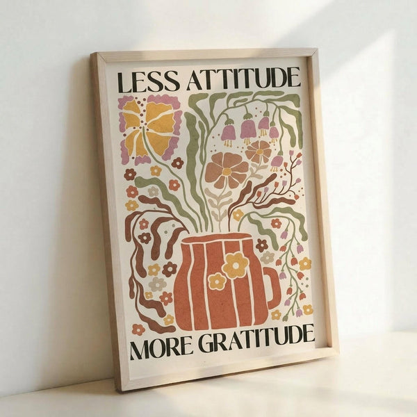 May include: Framed poster with colorful botanical illustration featuring a brown mug with flowers and vines flowing from it, including a yellow butterfly. Text reads "LESS ATTITUDE MORE GRATITUDE" in black letters. Artwork uses warm earth tones including orange, green, brown, and purple in a whimsical folk art style. Light wood frame sits against white wall on white surface.