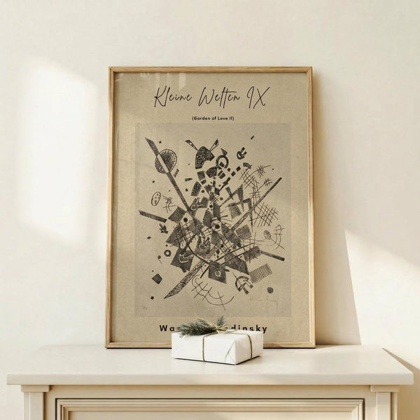 May include: Framed abstract art print in light wood frame displaying geometric black line drawings and shapes on beige background, leaning against white wall on light wooden surface beside small white gift box with twine and greenery accent.