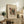 May include: Framed abstract art print displayed on rustic wooden sideboard in neutral-toned living room. Colorful geometric composition features red, yellow, teal, and black shapes with circles and patterns on cream background. Setting includes fern plant, ceramic vase, books, and beige upholstered chair with brown leather pillow.