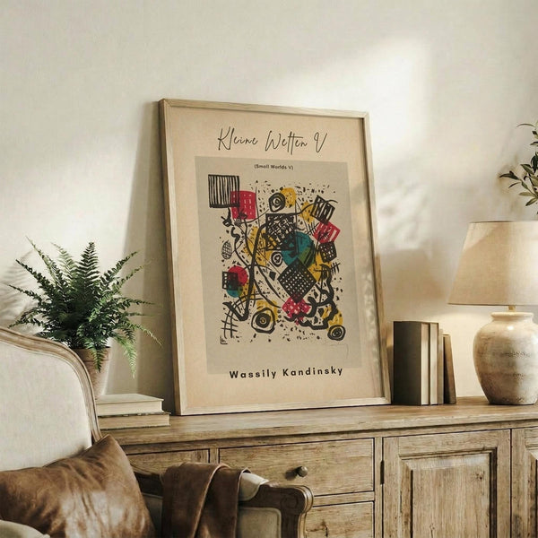 May include: Framed abstract art print displayed on rustic wooden sideboard in neutral-toned living room. Colorful geometric composition features red, yellow, teal, and black shapes with circles and patterns on cream background. Setting includes fern plant, ceramic vase, books, and beige upholstered chair with brown leather pillow.
