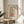 May include: Framed motivational poster with decorative floral design featuring a coffee mug with colorful botanical elements flowing from it, displayed on a round wooden side table next to a beige upholstered chair. Warm natural lighting creates soft shadows on white walls, with a plant visible in the corner of the cozy living room setting.