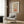 May include: Framed poster with retro-style illustration featuring colorful floral design in warm oranges, greens, and browns with text reading "Less Attitude More Gratitude." Displayed in modern living room setting with beige armchair, geometric patterned pillow, brass floor lamp, and wood-paneled wall. Natural lighting from window with white curtains.