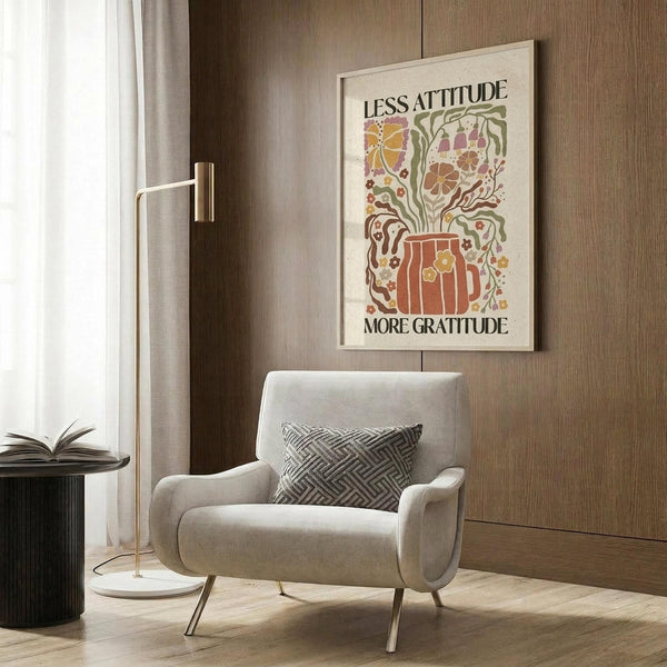 May include: Framed poster with retro-style illustration featuring colorful floral design in warm oranges, greens, and browns with text reading "Less Attitude More Gratitude." Displayed in modern living room setting with beige armchair, geometric patterned pillow, brass floor lamp, and wood-paneled wall. Natural lighting from window with white curtains.