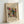 May include: Framed abstract art print in natural wood frame leaning against white wall. Colorful geometric composition with black lines, red squares, yellow shapes, teal circles, and various patterns including stripes and crosshatching. Artist signature "Wassily Kandinsky" visible at bottom. Modern minimalist display setting with soft lighting creating subtle shadows.
